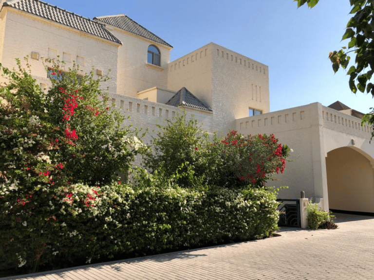 A large beige villa with arched entrances surrounded by lush green bushes and red flowering plants under a clear blue sky.