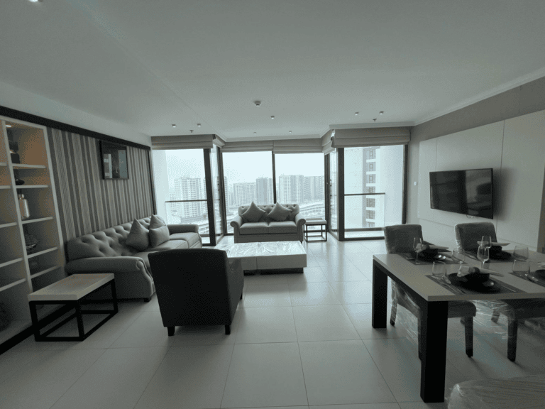 Modern 2 BR apartment living room with gray sofas, a tv, and a dining area, leading to a balcony with city views.