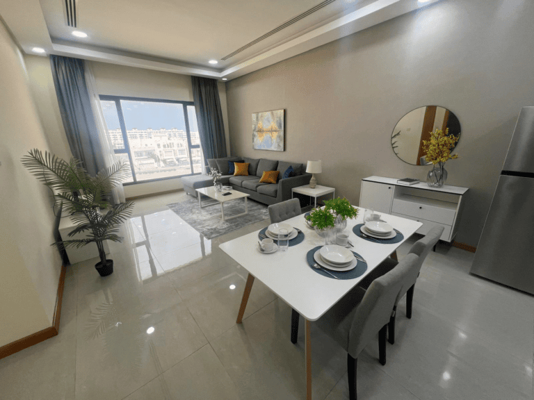 Modern apartment living room with a dining area; features a grey sofa, dining table set for four, and large windows with city view. Decor includes plants and minimalist artwork.