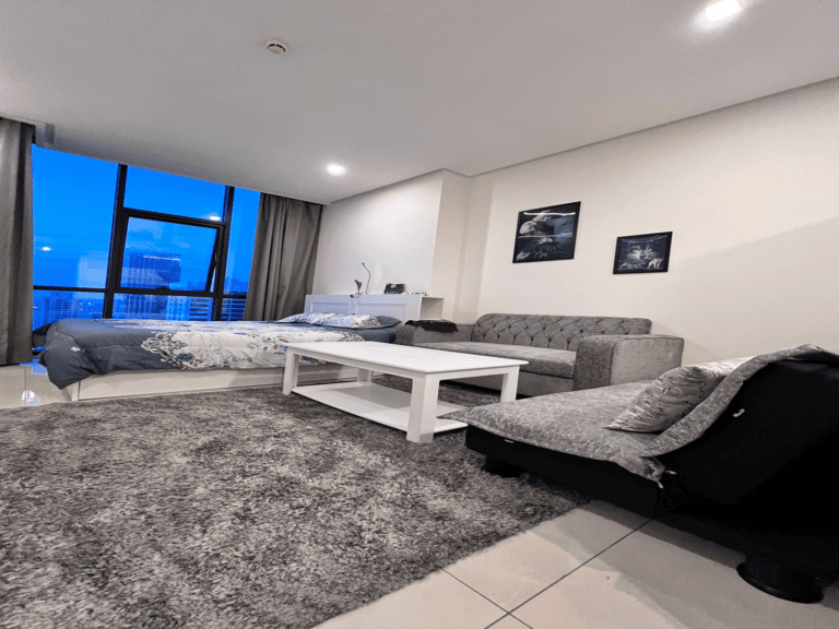 Modern living room with a gray sofa, white coffee table, large bed, and wall art, featuring a balcony entrance with city views.