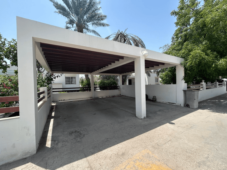 A 3 BDR Villa in the Hamala Area featuring a carport with a white structure and wooden slats for a roof, surrounded by greenery and palm trees on a sunny day.