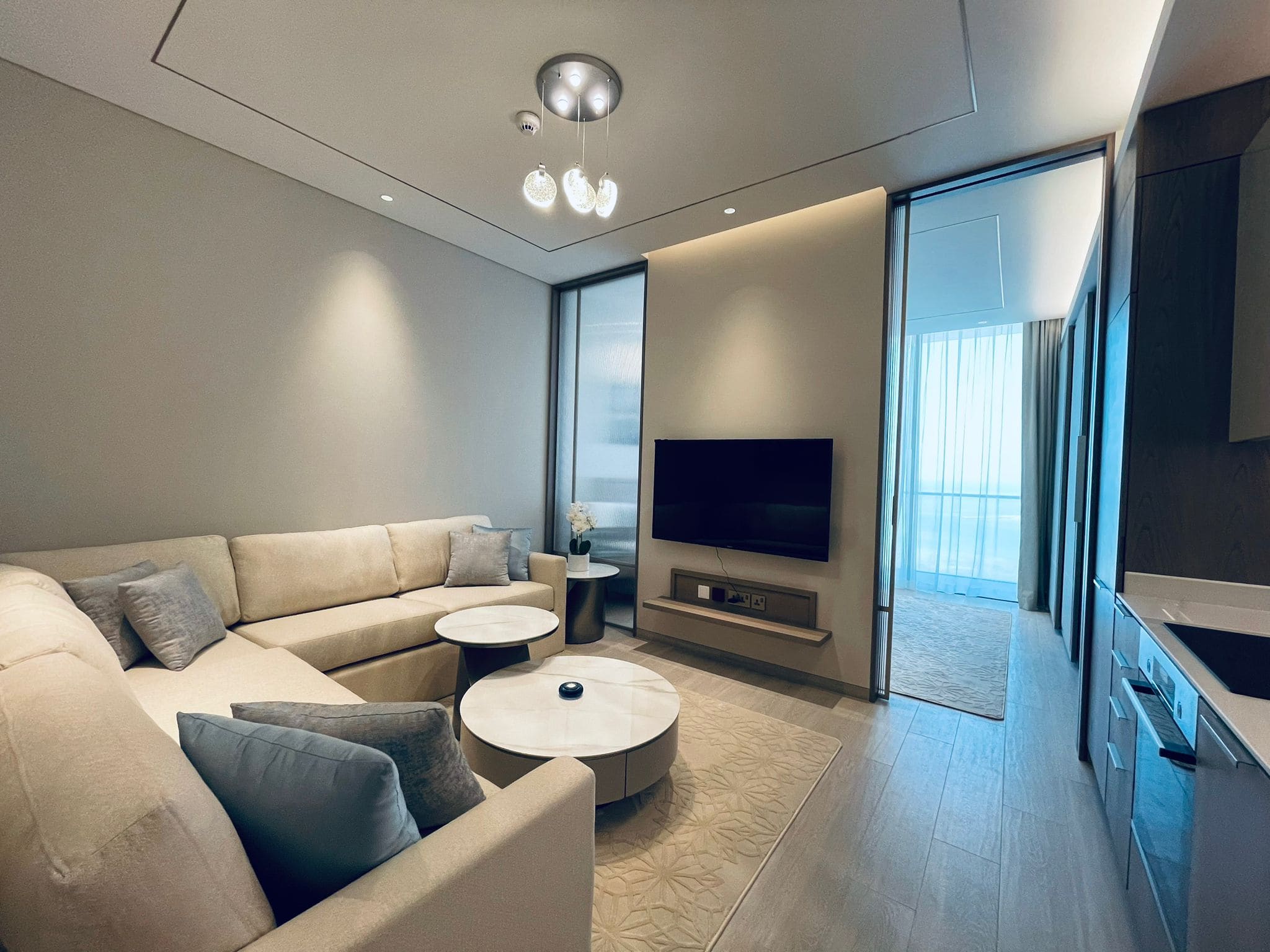 Modern living room in an amazing apartment for rent in Seef, featuring a beige sectional sofa, round coffee table, flat-screen TV, and sleek cabinetry, with ample natural light filtering through sheer curtains