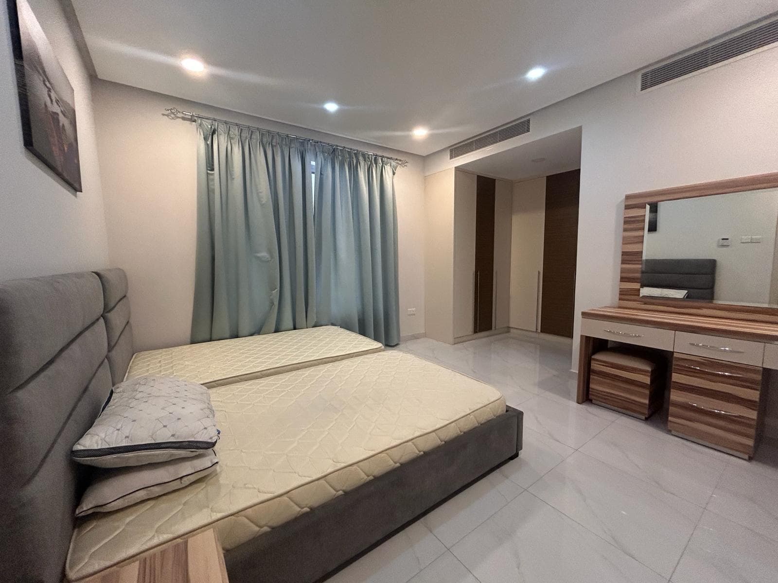 A modern bedroom in a luxury apartment with a gray upholstered bed, wooden furniture, and pale curtains.