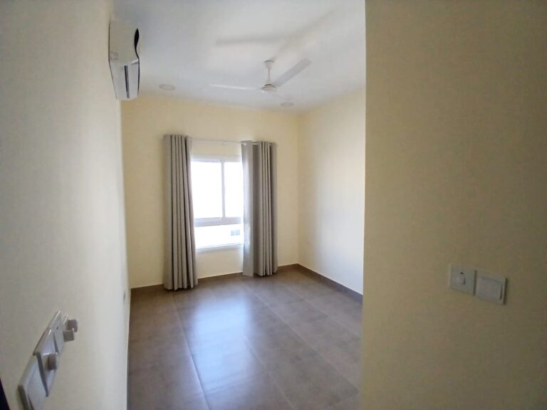 An empty room in a brand new apartment with beige walls, a ceiling fan, tiled floor, and a window with curtains.