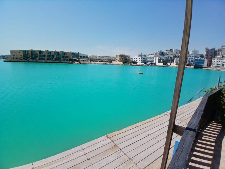 Coastal urban landscape viewed from a wooden pier in Amwaj, featuring turquoise waters and modern buildings along the shore, including luxurious 3BR Seafront Villas available for rent.