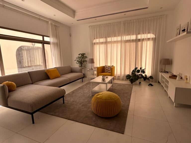 غرفة معيشة حديثة بألوان محايدة، تحتوي على أريكة رمادية مع وسائد صفراء، وطاولة قهوة بسطح زجاجي، ومسند للأريكة باللون الأصفر.