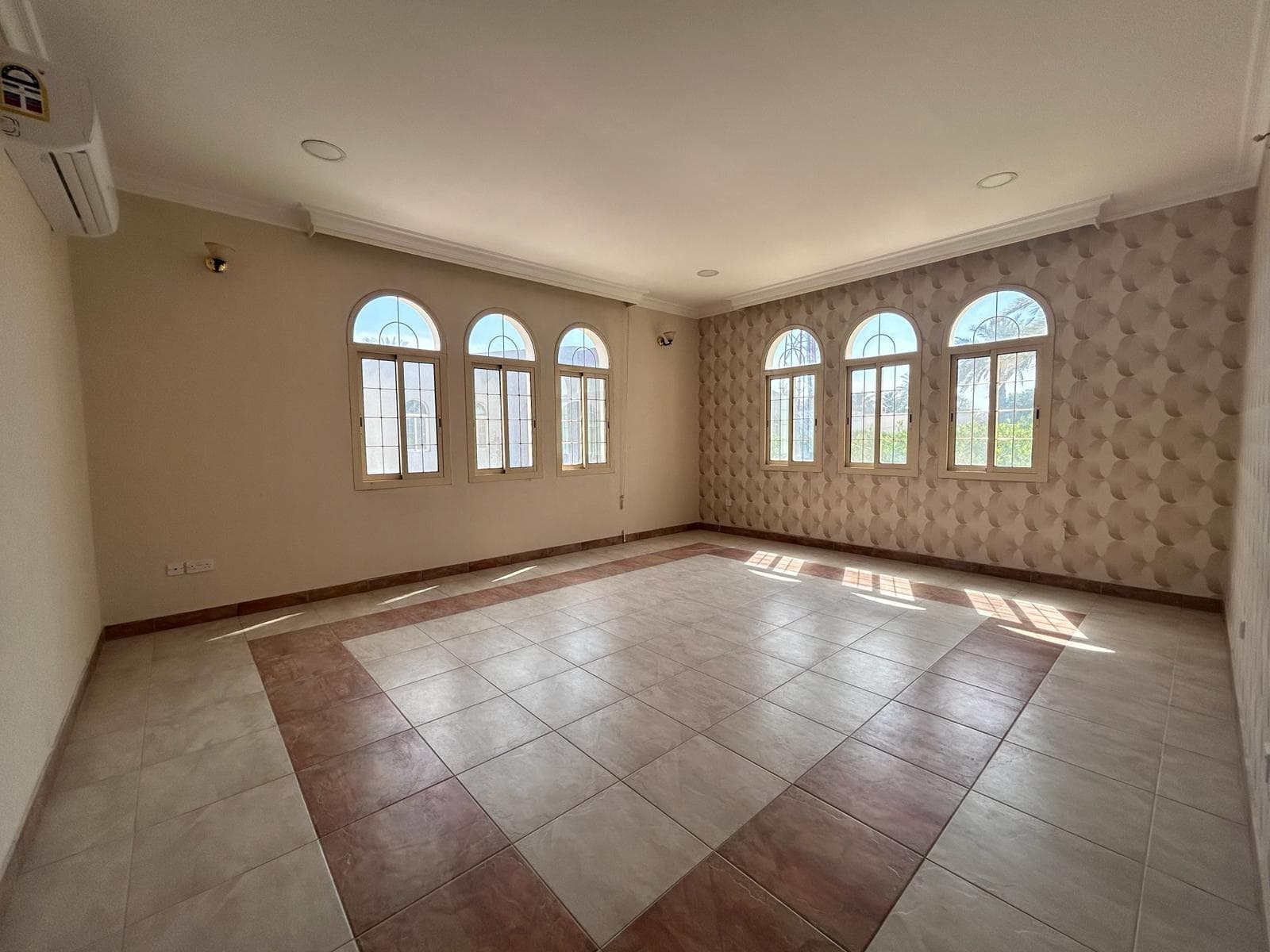 Empty living room with beige walls, arched windows, and tiled floor, sunlight streaming in, creating patterns on the floor.