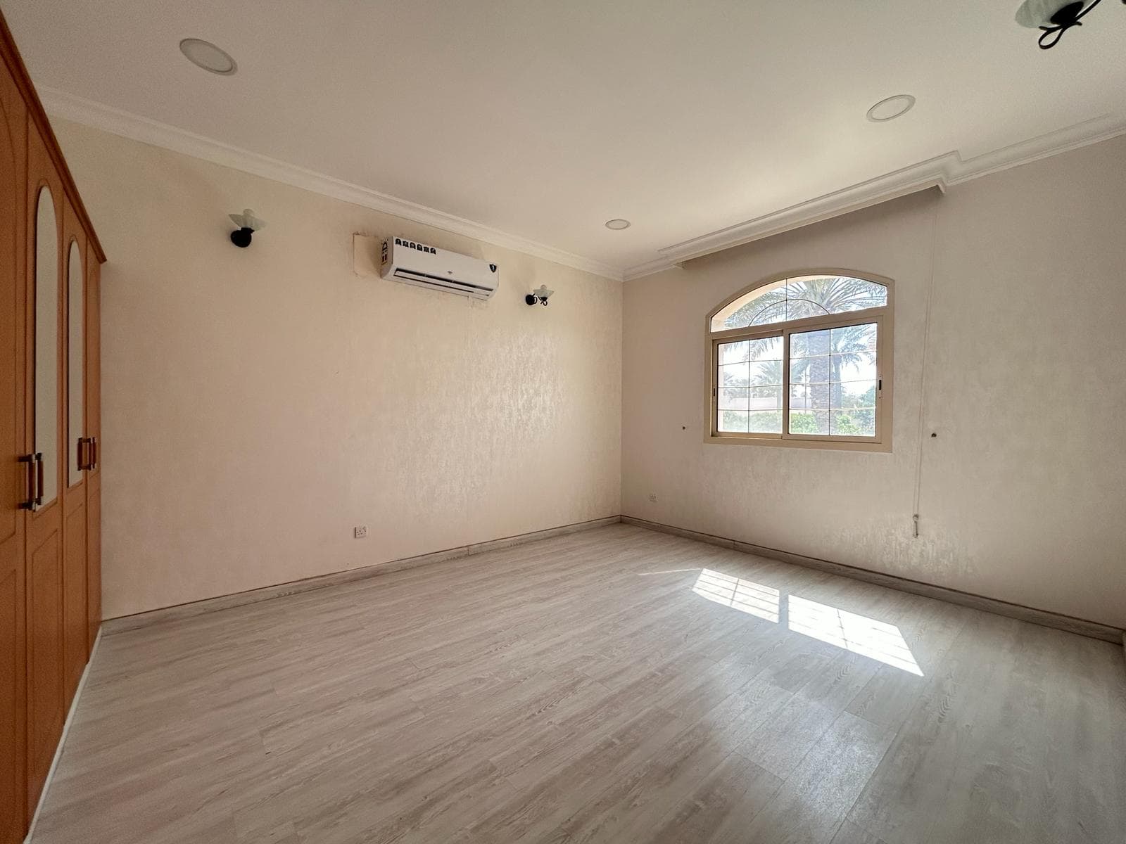 Empty room with beige walls, wooden flooring, arched window, and an air conditioner unit mounted on the wall.