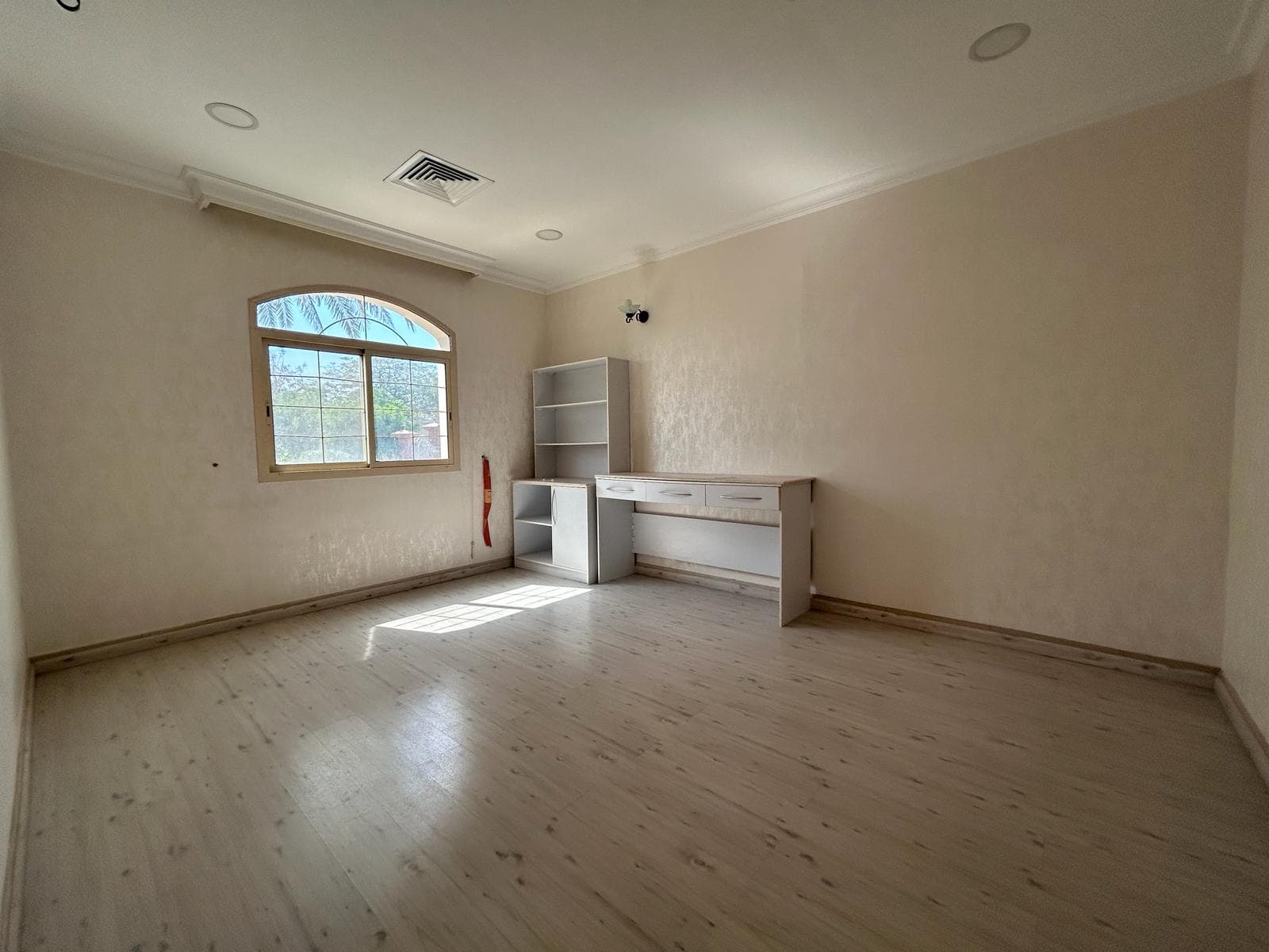 Empty room with pale walls and light wooden flooring, featuring an arched window and a built-in white cabinet.