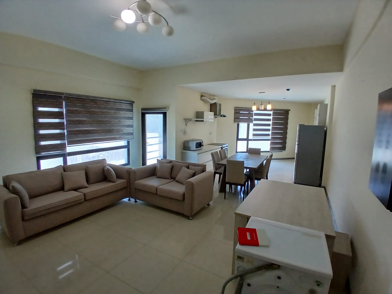 Modern living room with beige sofas, a dining area, and an open kitchen in Al Hidd Area, illuminated by natural light and stylish ceiling lights.