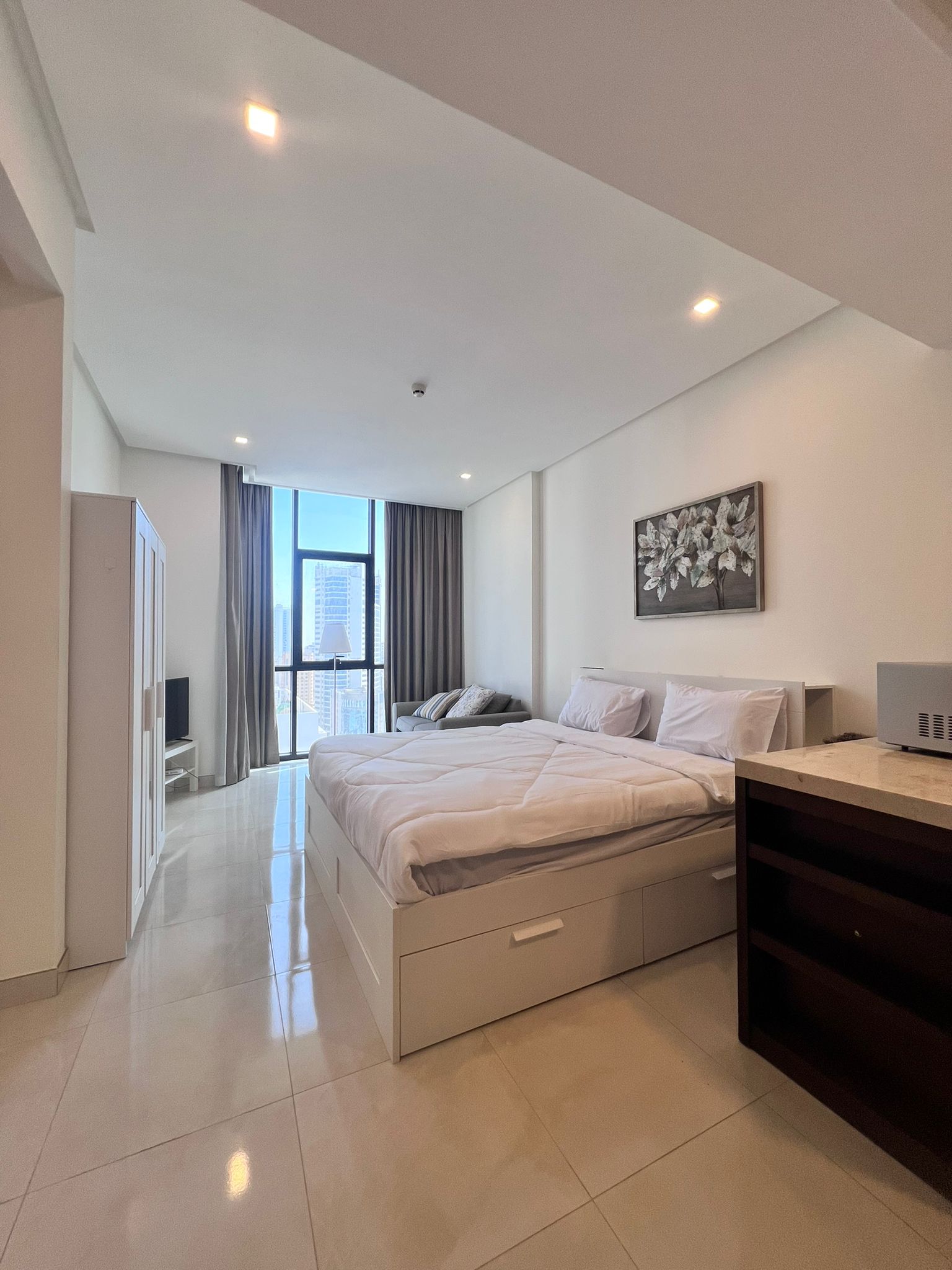 Modern bedroom in a Juffair flat for sale, featuring a large bed, white bedding, a painting of flowers above, and a view of the city through a window.