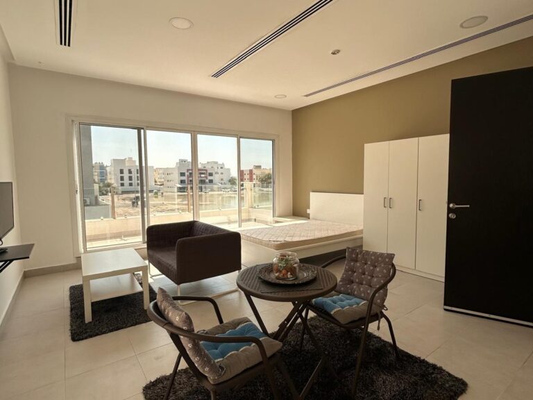 غرفة معيشة شقة حديثة مع نافذة كبيرة تطل على المناظر الطبيعية الحضرية، وتحتوي على أريكة بنية وكرسيين وطاولة قهوة وخزائن بيضاء.