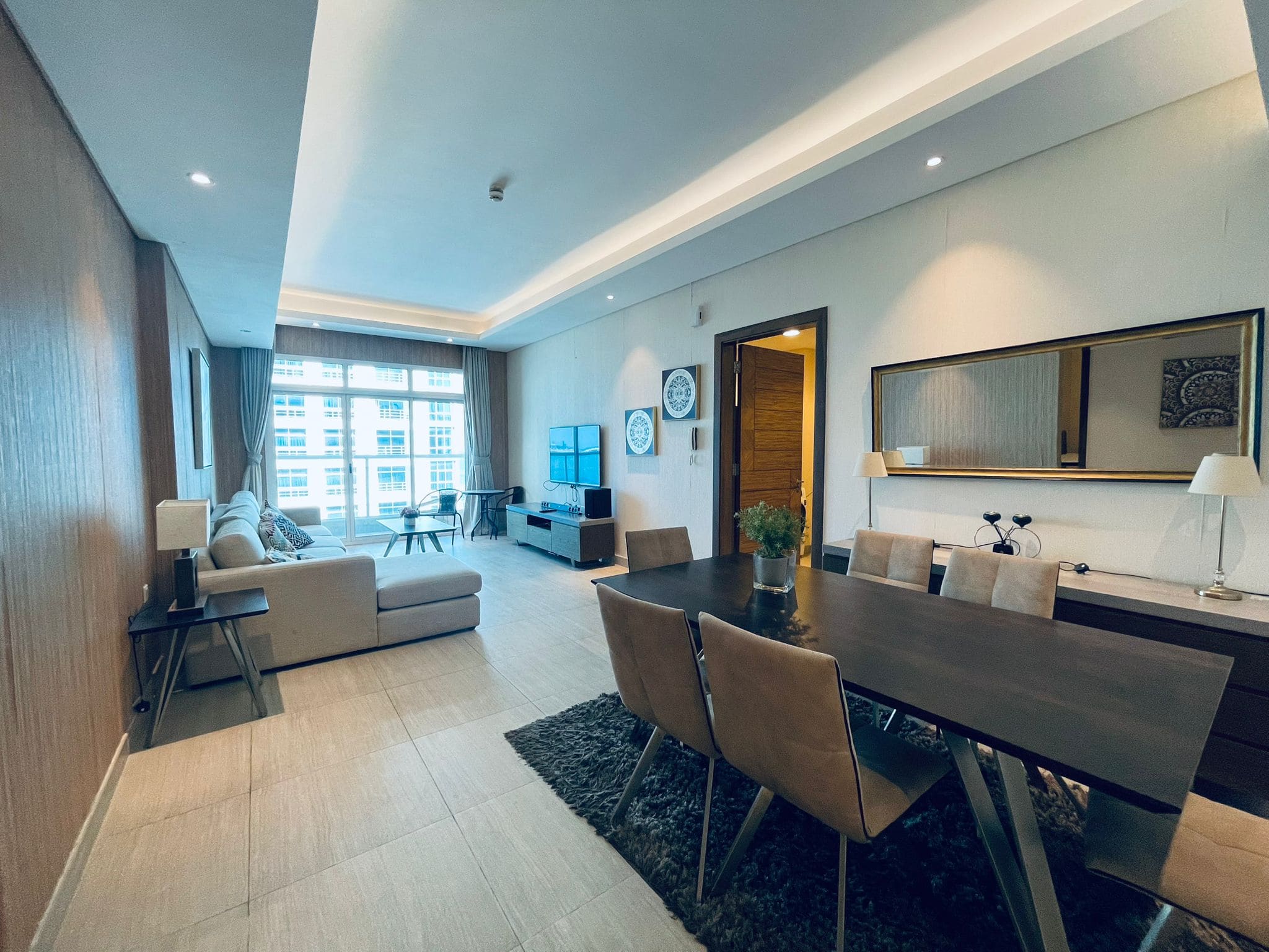 Modern apartment living room with a dining area, featuring a beige couch, wooden dining table, and views of the city through large windows.