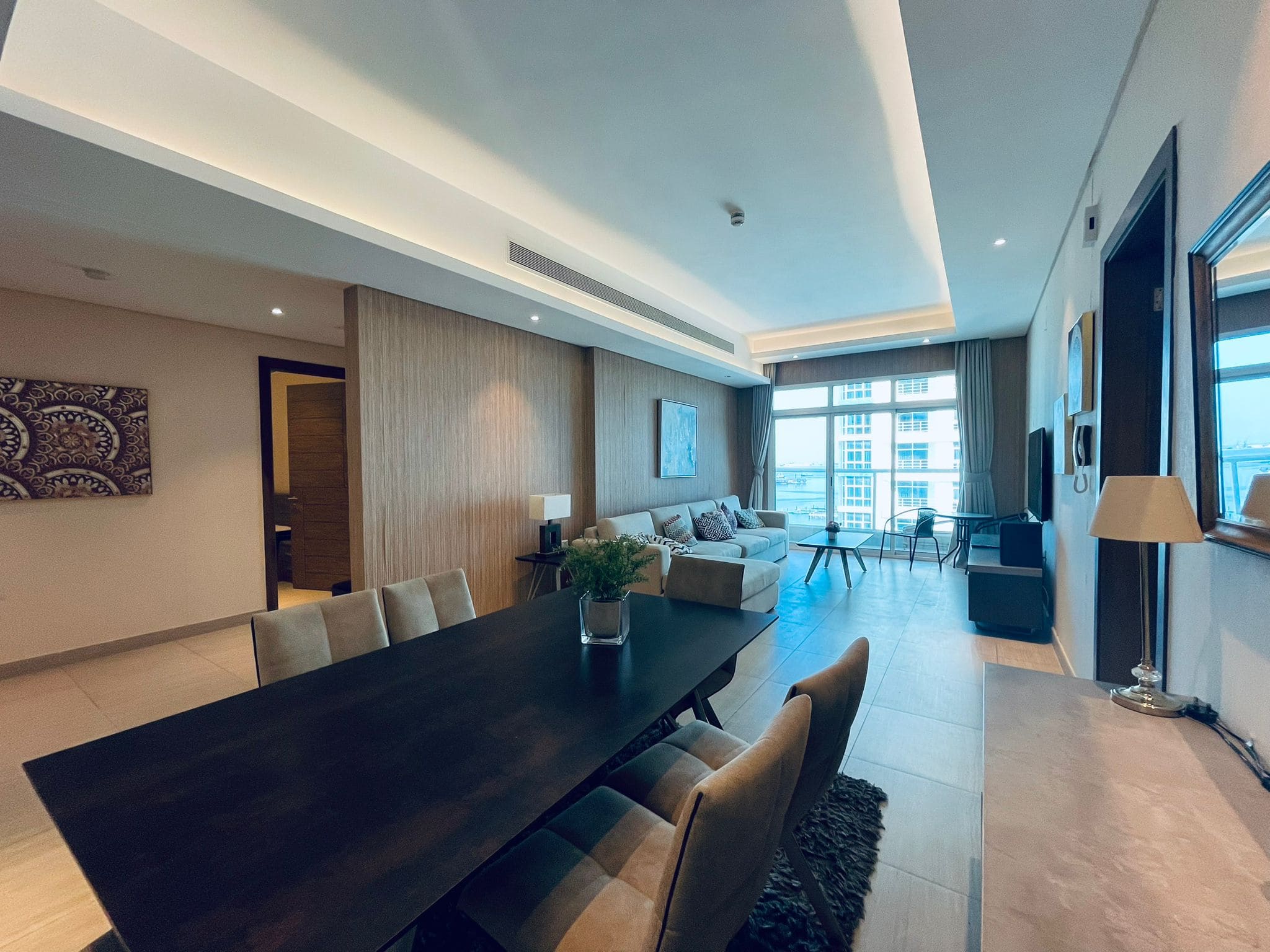 Modern apartment interior featuring a long dining table, a cozy living area with a sofa, and large windows providing ample auto draft and natural light.