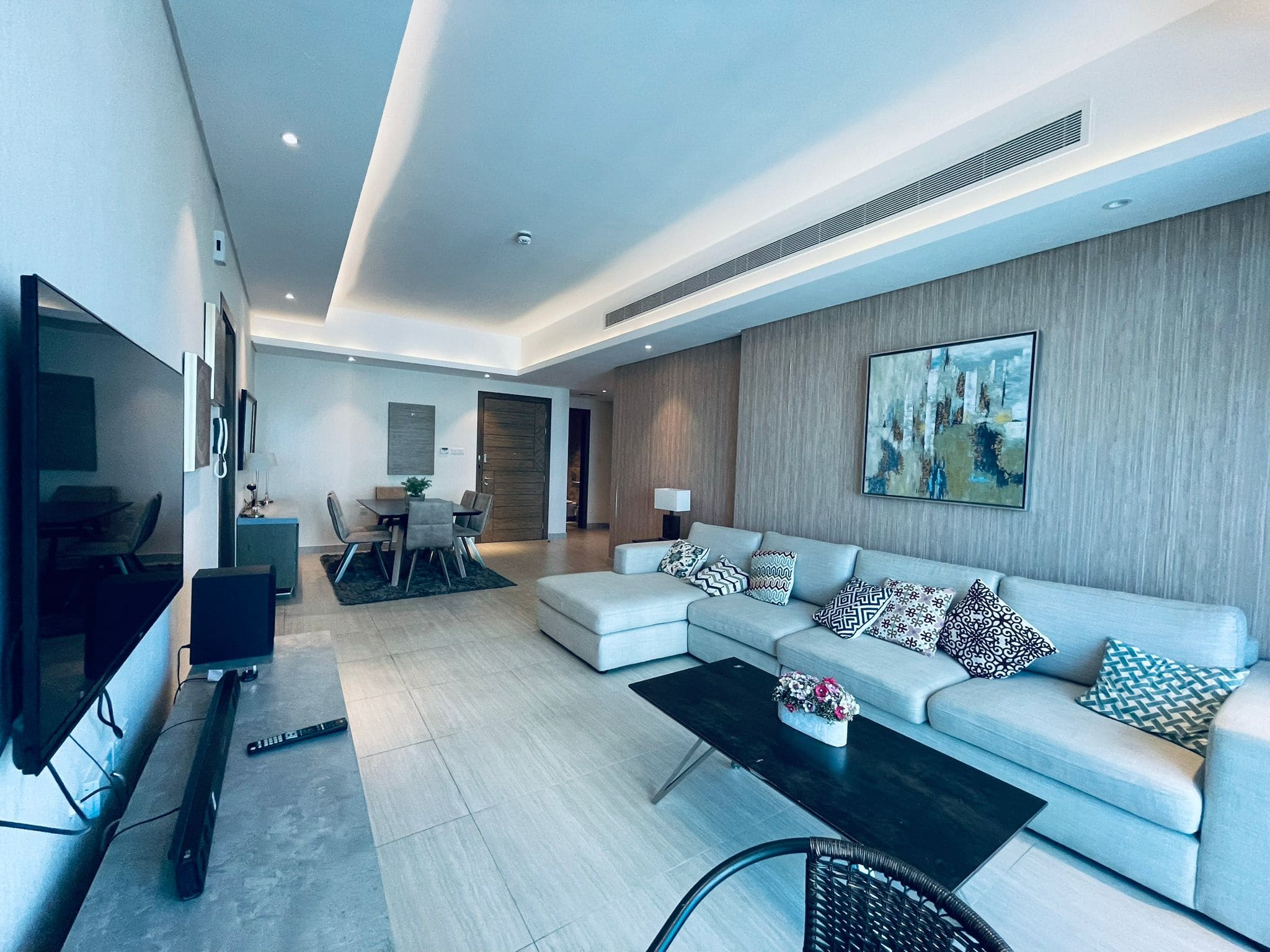 Modern living room with a light gray sectional sofa, patterned cushions, a glass coffee table, and a large wall-mounted TV featuring neutral colors and auto draft.