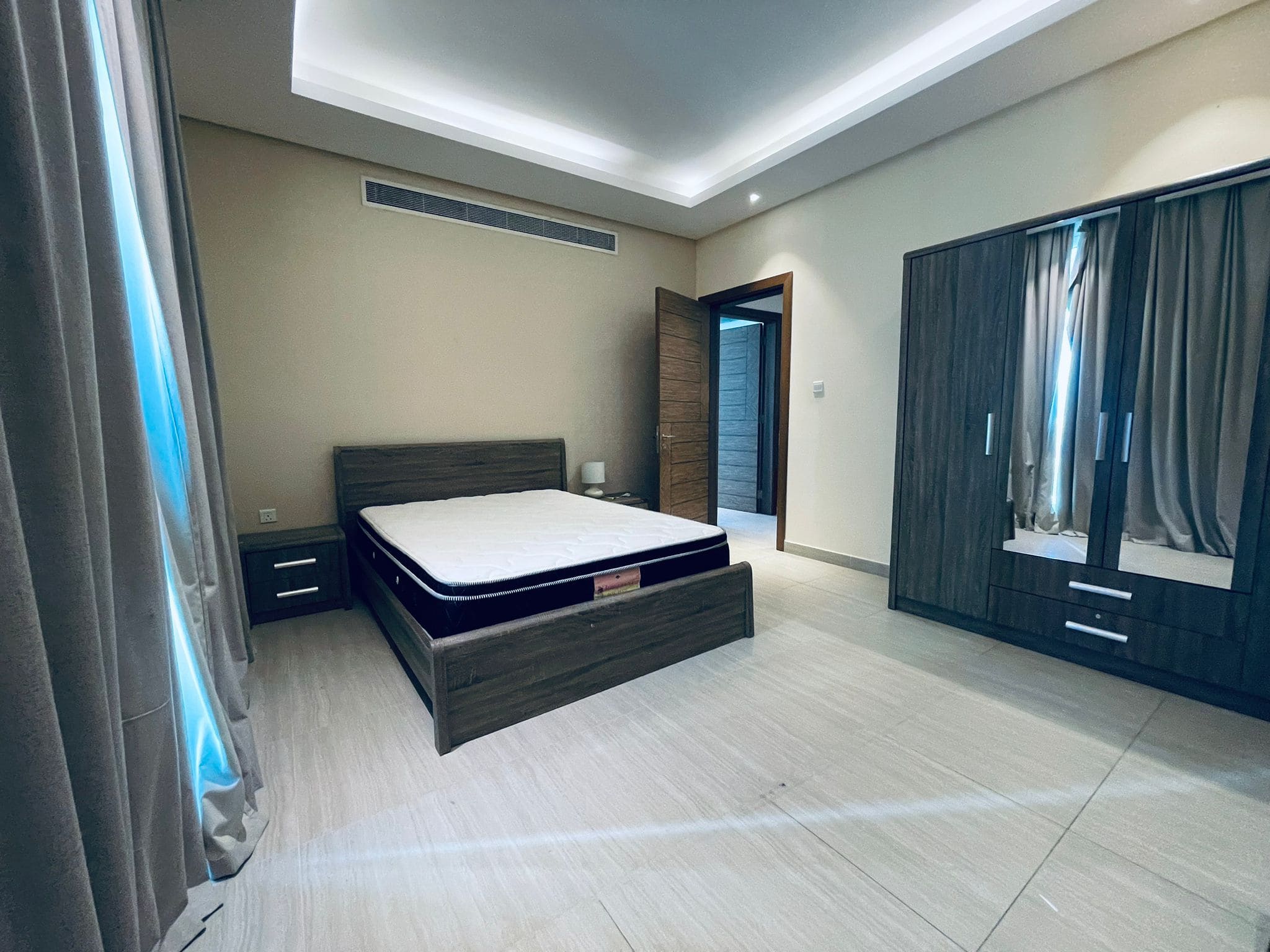 Modern bedroom featuring a large bed with a dark headboard, wooden wardrobes, and Auto Draft tiled flooring, with subtle grey and blue curtains.