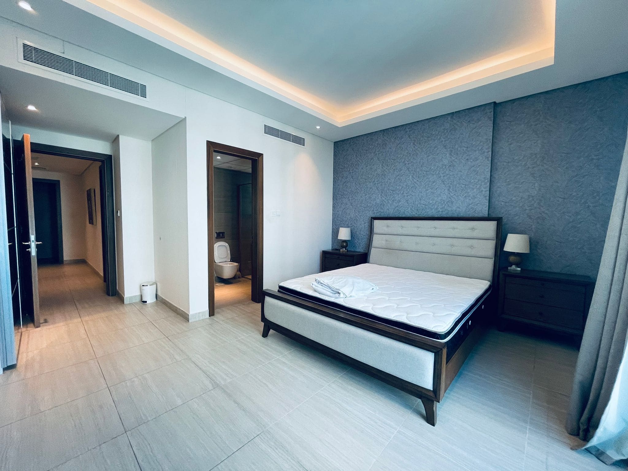 Modern bedroom with a large bed, blue accent wall, ambient lighting, and an open door leading to an auto draft bathroom.