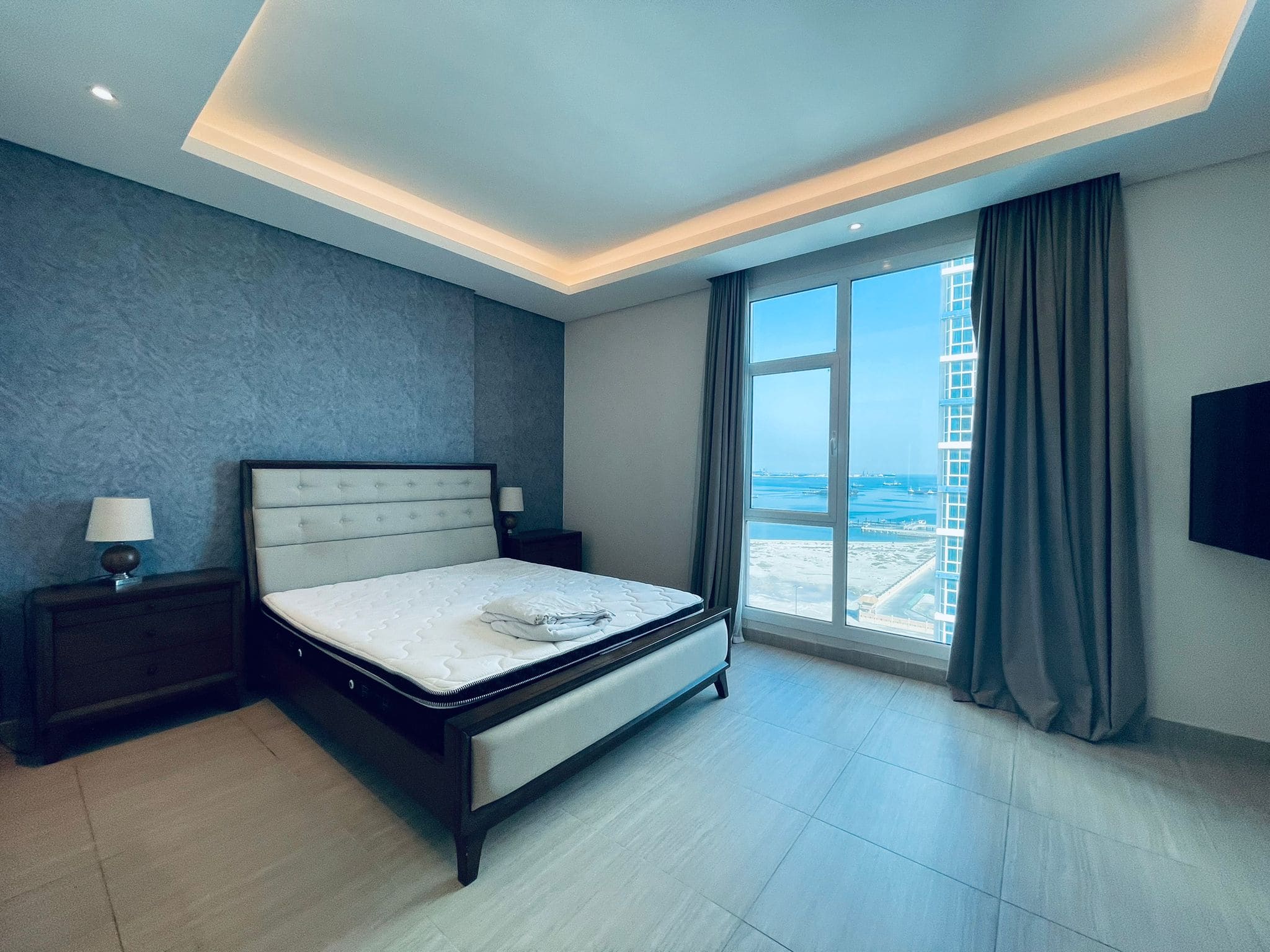 Modern bedroom with a large bed, side tables, and a floor-to-ceiling window overlooking an auto-drafted coastal cityscape.