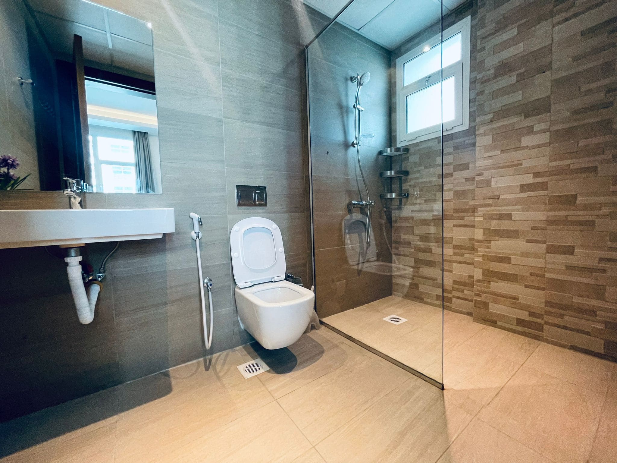 Modern bathroom interior with a wall-mounted toilet, glass shower area, and beige tiled walls. A mirror and sink are visible near a window. Auto Draft