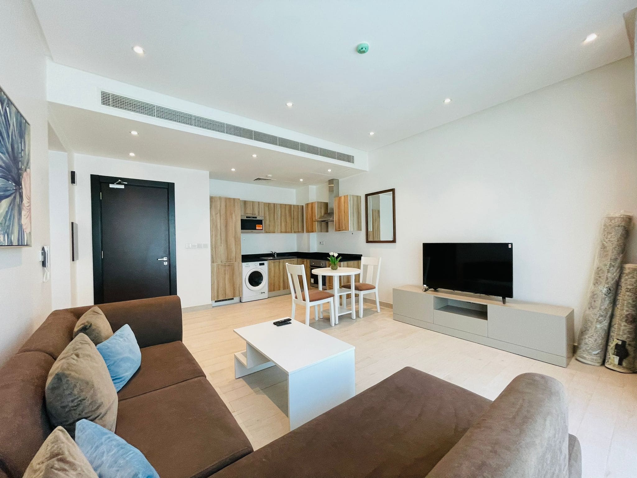 Stylish apartment interior showing a living room with a brown sofa, white coffee table, and an integrated kitchen area with appliances.