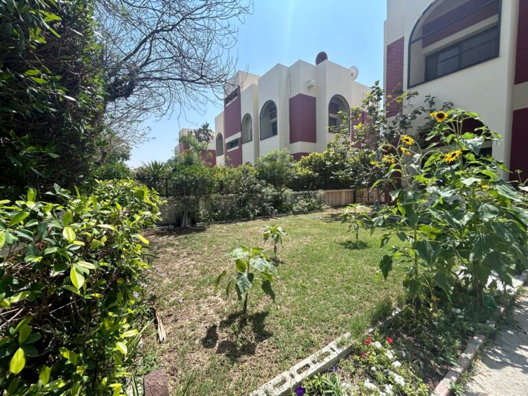 حديقة مورقة بها عباد الشمس ونباتات متنوعة أمام مبنى ذو هندسة معمارية مغربية باللونين الوردي والأبيض تحت سماء صافية.