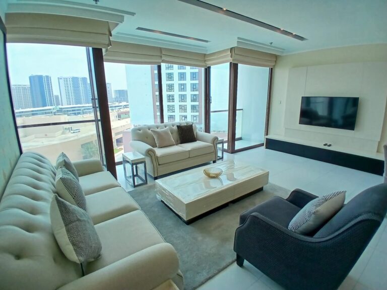 غرفة معيشة حديثة ذات نوافذ كبيرة توفر إطلالات على المدينة، وتحتوي على أريكتين وكراسي بذراعين وطاولة قهوة مركزية.