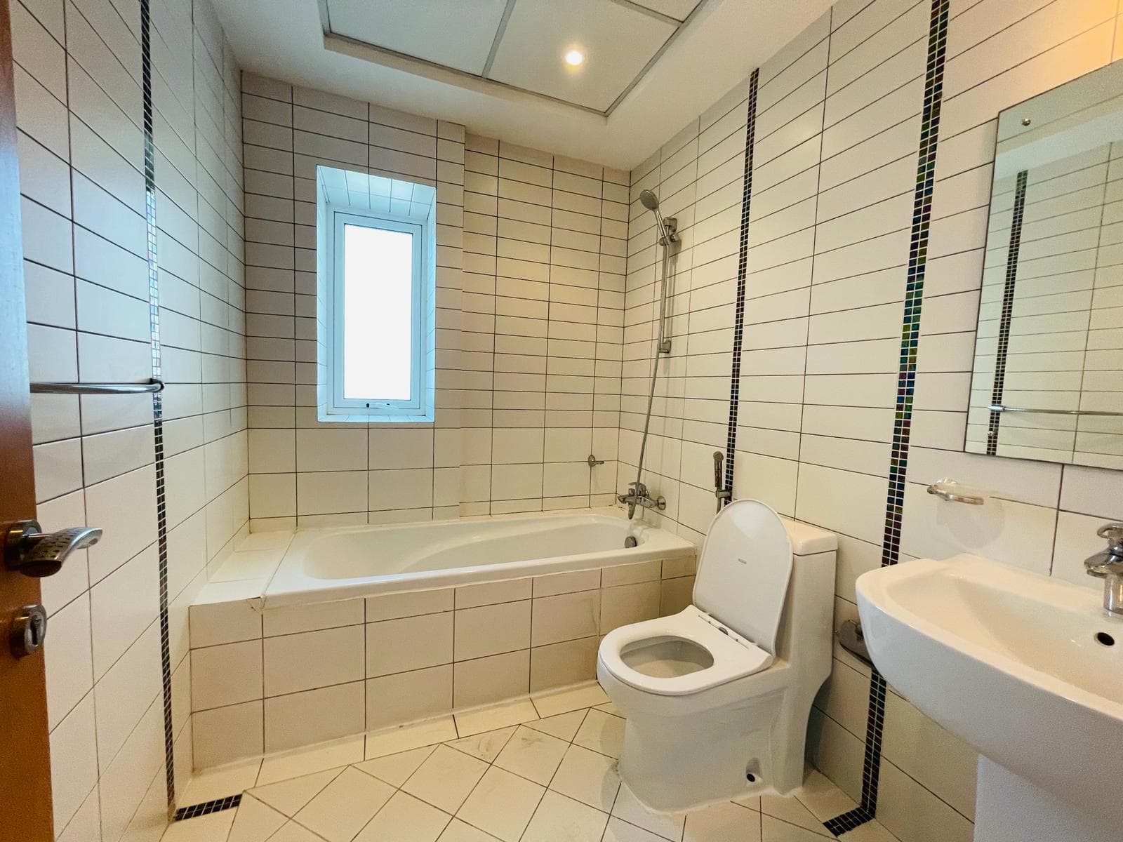 Luxury apartment bathroom with a white bathtub, toilet, and sink, tiled walls, and a small window letting in natural light.