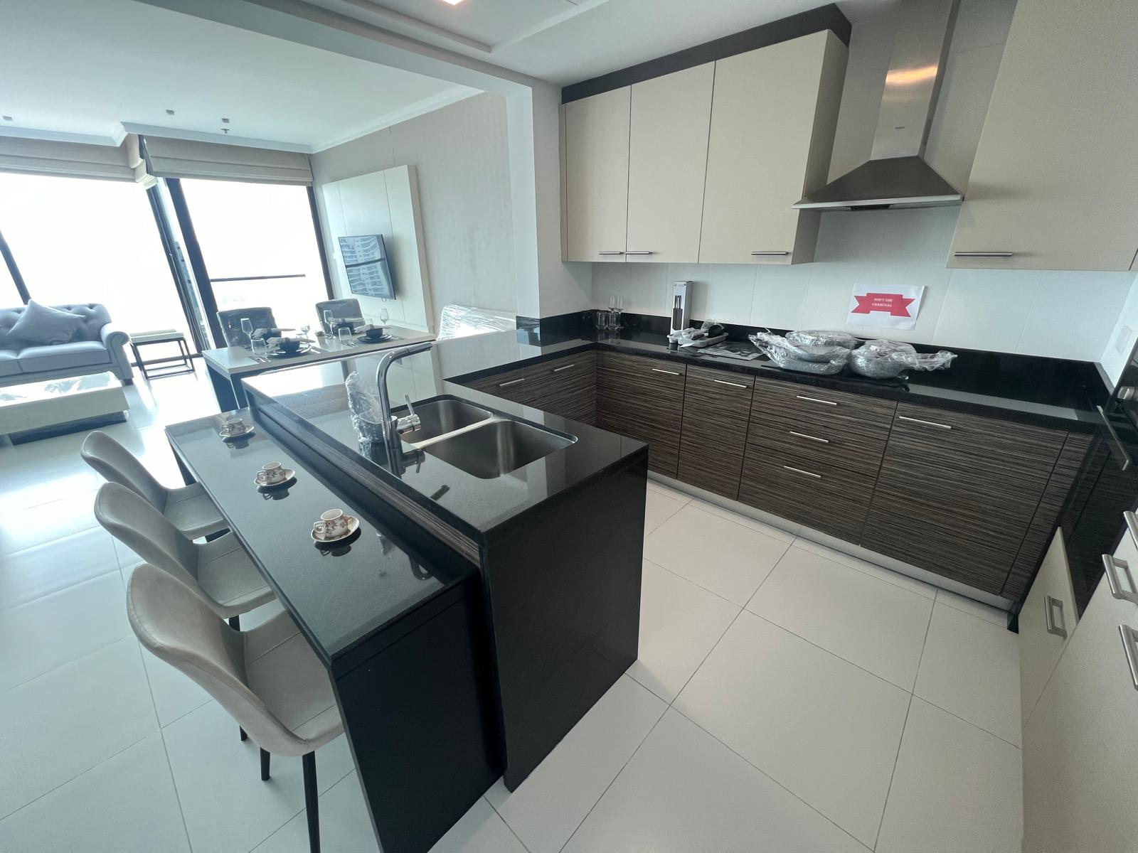 Modern kitchen interior in a fully furnished 2 BR apartment featuring a black island counter, bar stools, built-in cabinets, and stainless steel appliances, with natural light from adjacent windows.