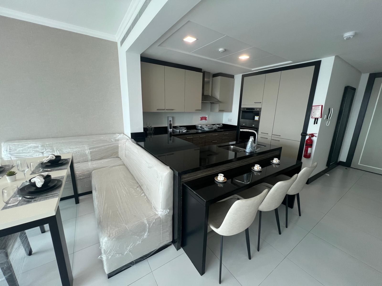 Modern kitchen in a fully furnished 2 BR apartment with white cabinets and black countertops, including an island and dining area with chairs still wrapped in protective plastic.