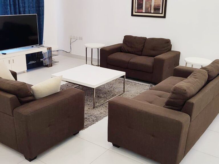غرفة معيشة مرتبة بشكل أنيق وتحتوي على طقم أريكة بني، وطاولة قهوة بيضاء، وتلفزيون.