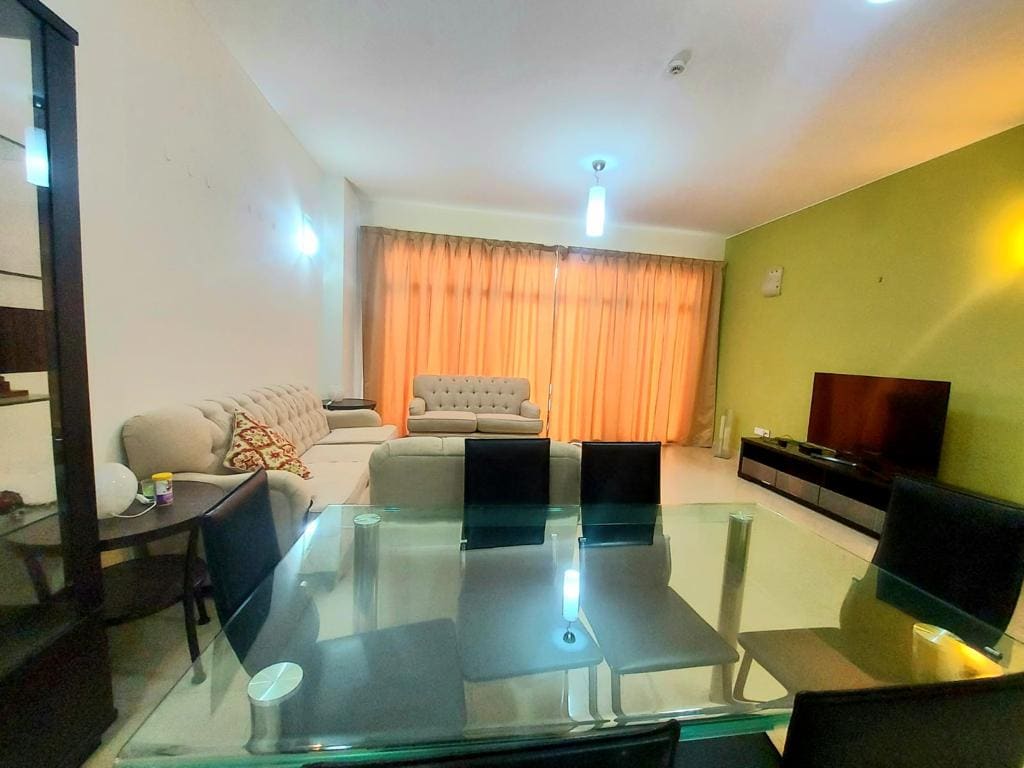 Spacious flat for sale in Amwaj with a living room featuring a gray sofa, matching loveseat, glass dining table with four chairs, and a television unit, illuminated by ceiling lights, with orange curtains.