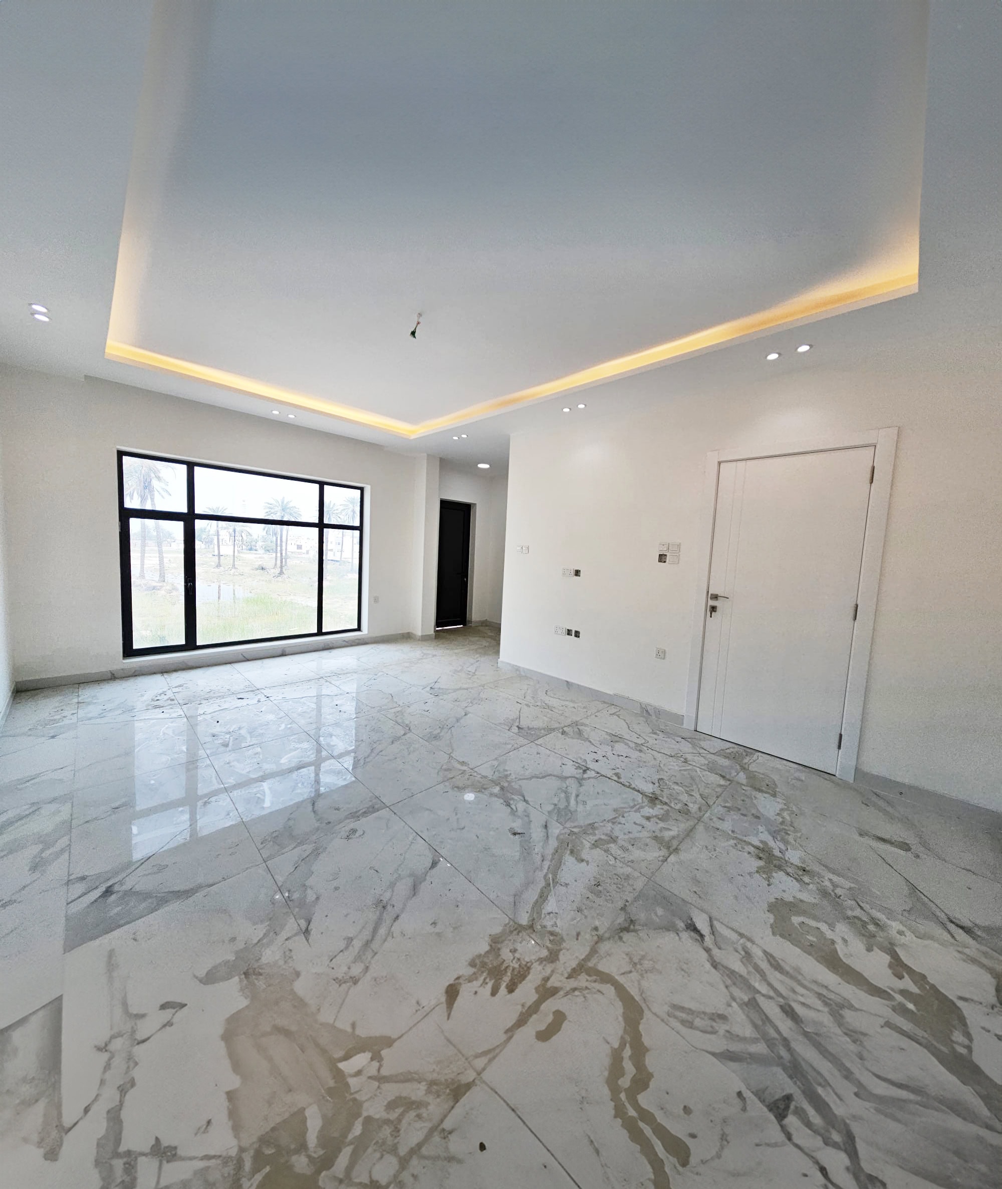 A luxurious villa boasts a modern, empty room with marble flooring, white walls, a large window, and a single door. The ceiling features recessed lighting. Currently on sale, this elegant space offers the perfect blend of sophistication and comfort.