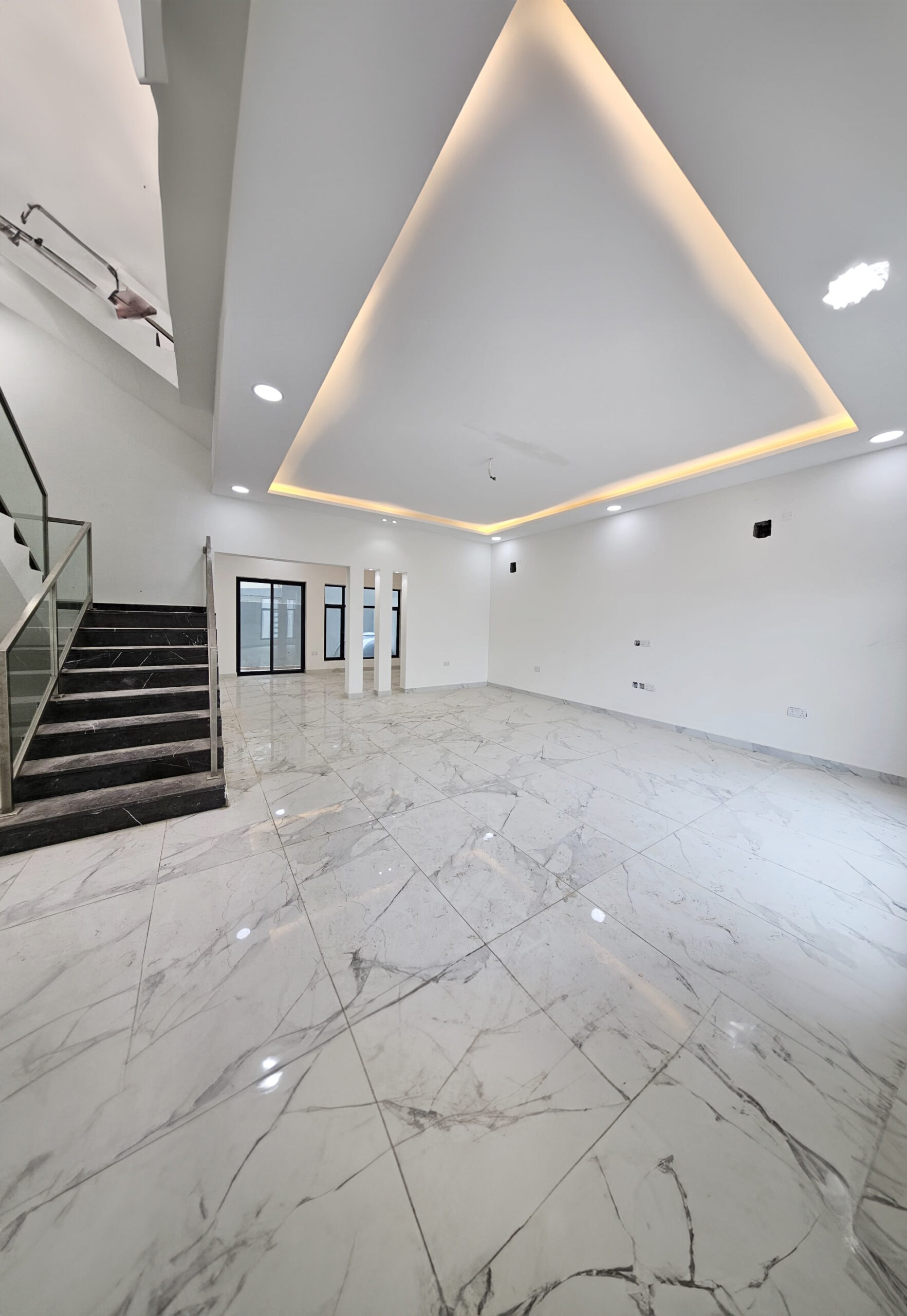 A luxurious, modern villa room featuring marble floors, a staircase with glass railing, recessed ceiling lights, and a large ceiling cut-out with soft lighting. Walls are white with light fixtures and outlets.