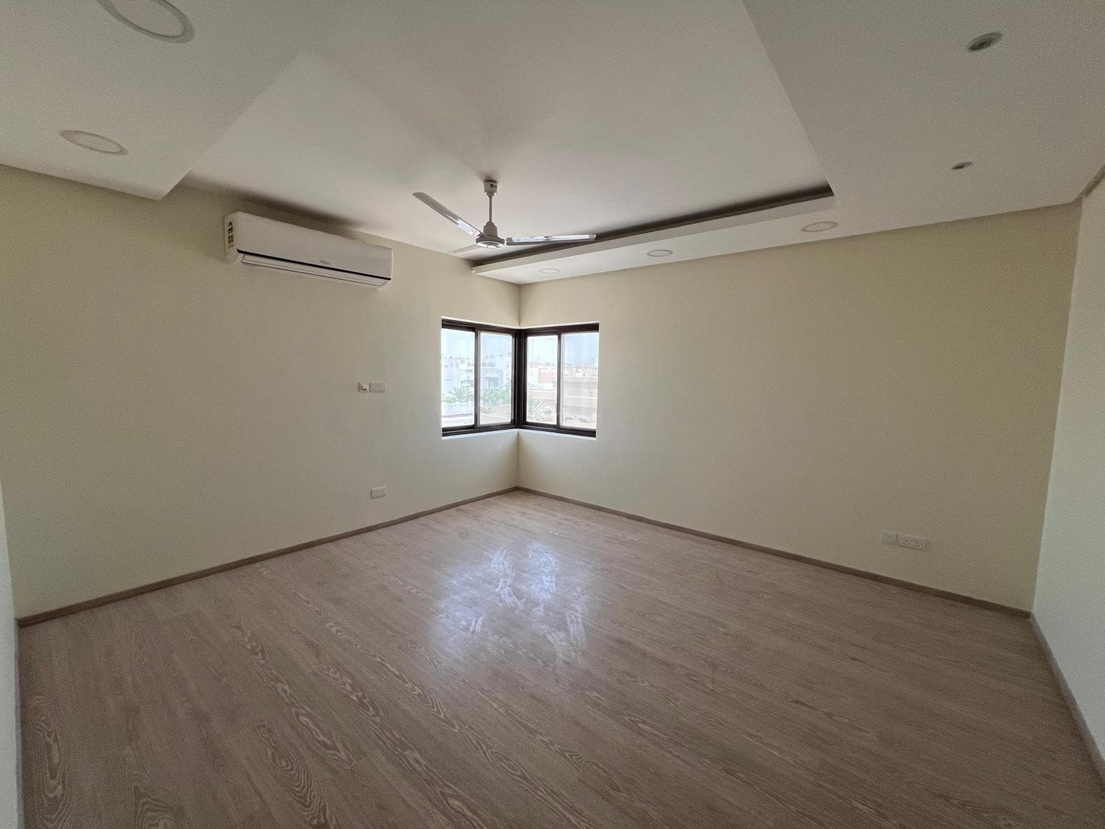 This beautiful villa in Sanad is for rent, featuring an empty room with light wood flooring, a ceiling fan, and an air conditioning unit mounted on the wall. The room has recessed lighting and a large window allowing natural light to enter.