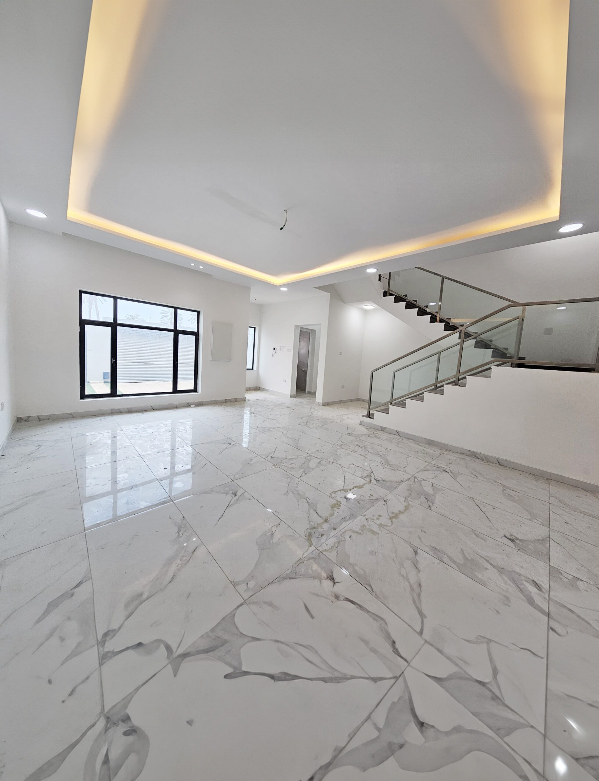 A spacious, bright room with white marble flooring, a staircase with glass railing, a large window, and recessed ceiling lighting in this luxurious villa for sale in Barbar.