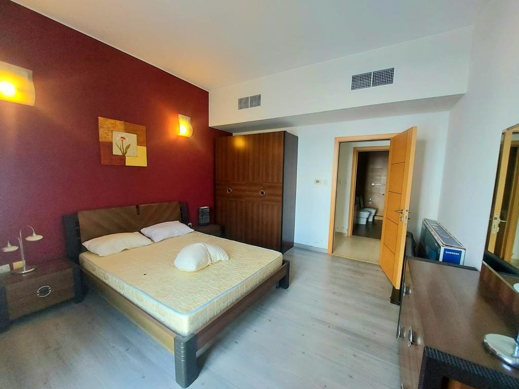 A well-lit flat in Amwaj featuring a large bed with white bedding against a deep red wall, wooden flooring, an open door, and minimalistic decor.