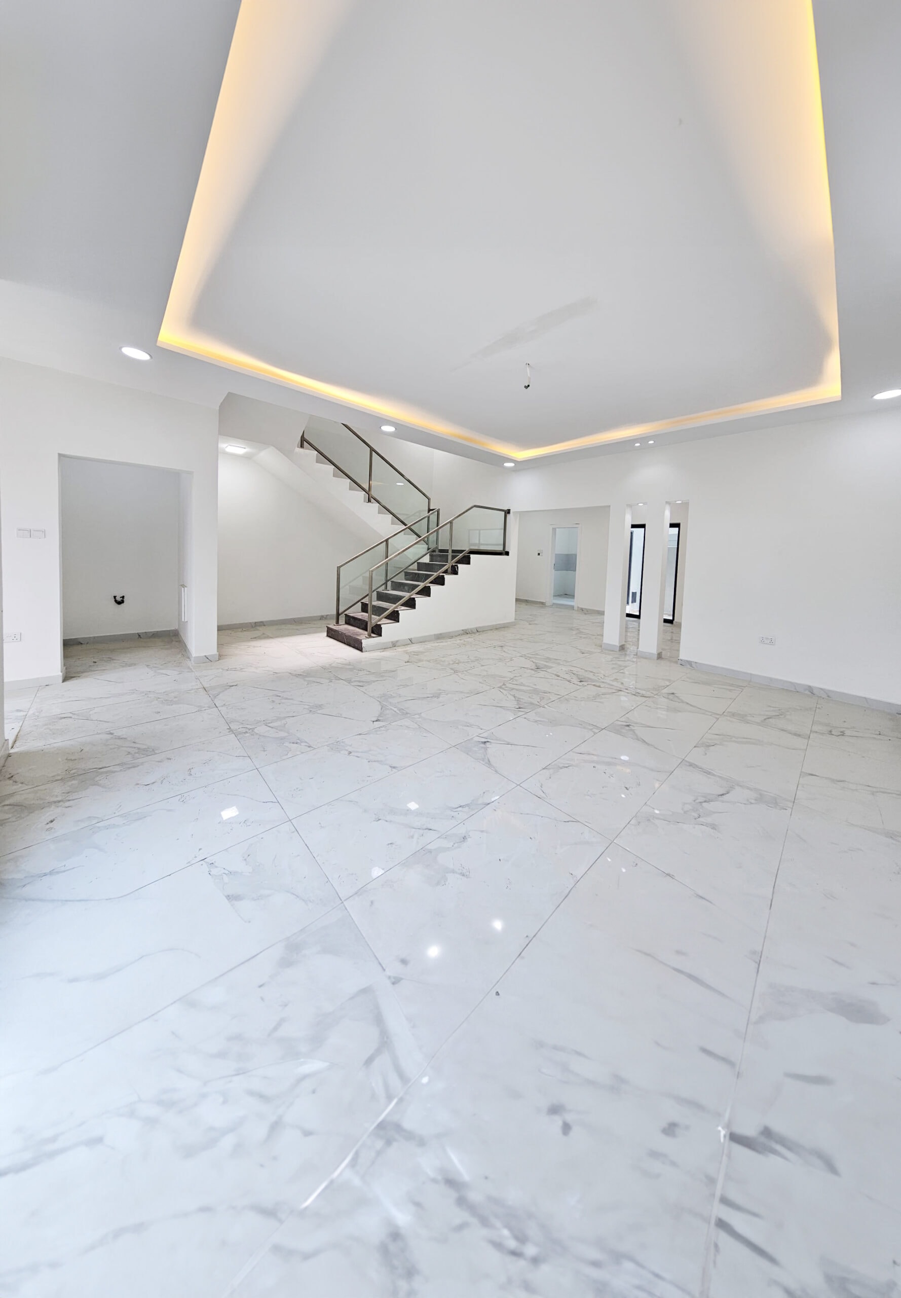 A modern, spacious room in a luxurious villa with white marble flooring, a staircase with a glass railing, recessed lighting in the ceiling, and white walls. Two open doorways lead to adjacent rooms in this stunning property for sale.