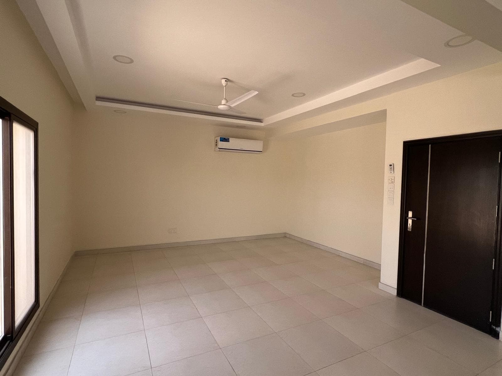 An empty room in a Sanad villa awaits, featuring tiled flooring, a ceiling fan, mounted air conditioning unit, large window, and a dark wooden door. Perfect for rent!