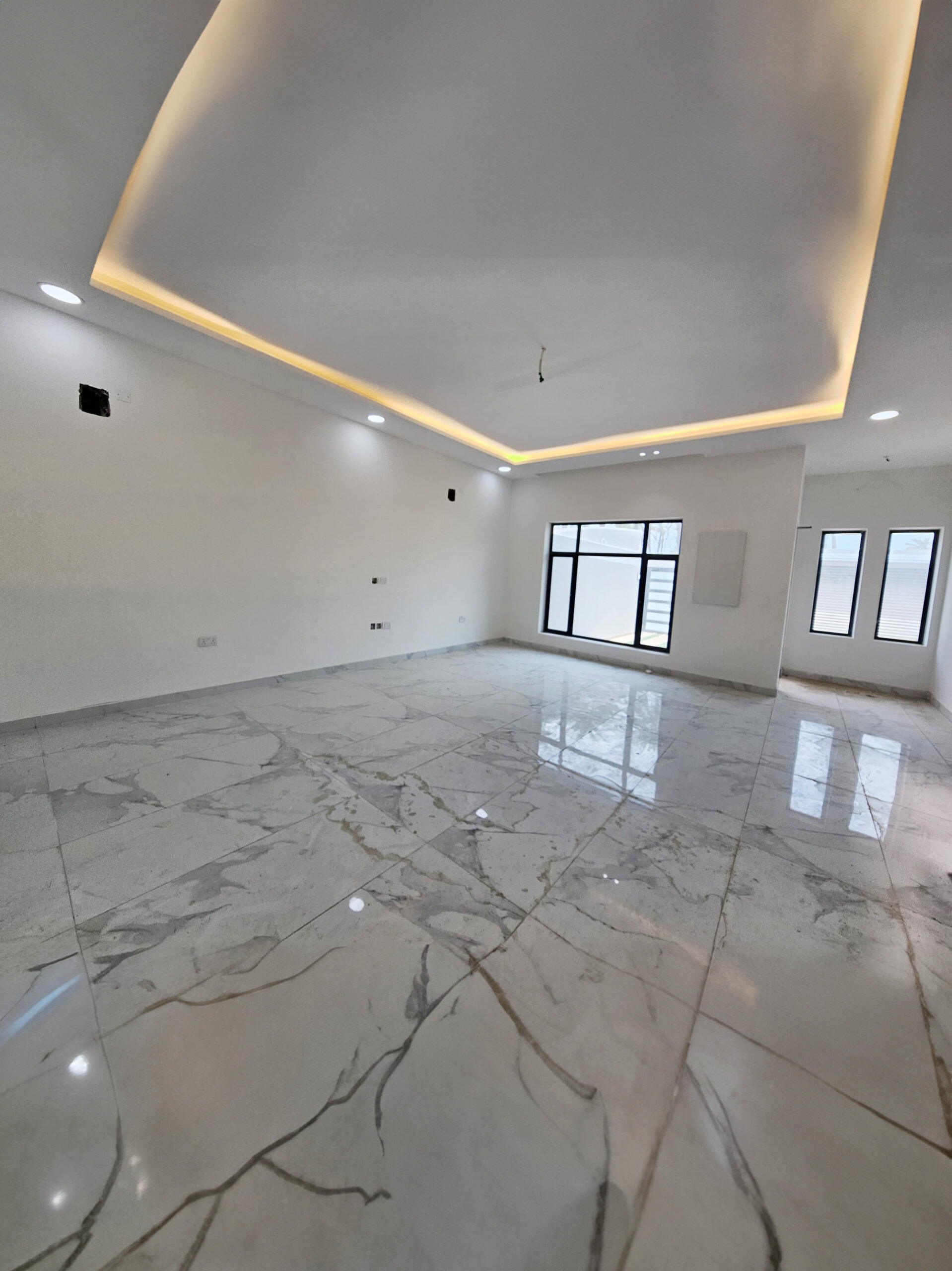 A spacious room in a luxurious villa with a white ceiling featuring recessed lighting, marble flooring, and large windows letting in natural light.