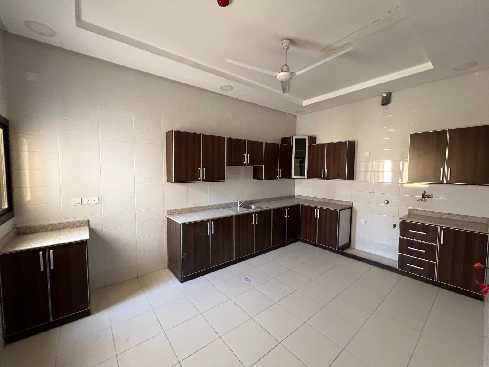 A spacious kitchen in the beautiful Sanad villa features brown wooden cabinets, a white tile backsplash, granite countertops, a single sink, and white tile flooring. There is a ceiling fan and a window with natural light. This villa for rent offers both elegance and comfort.