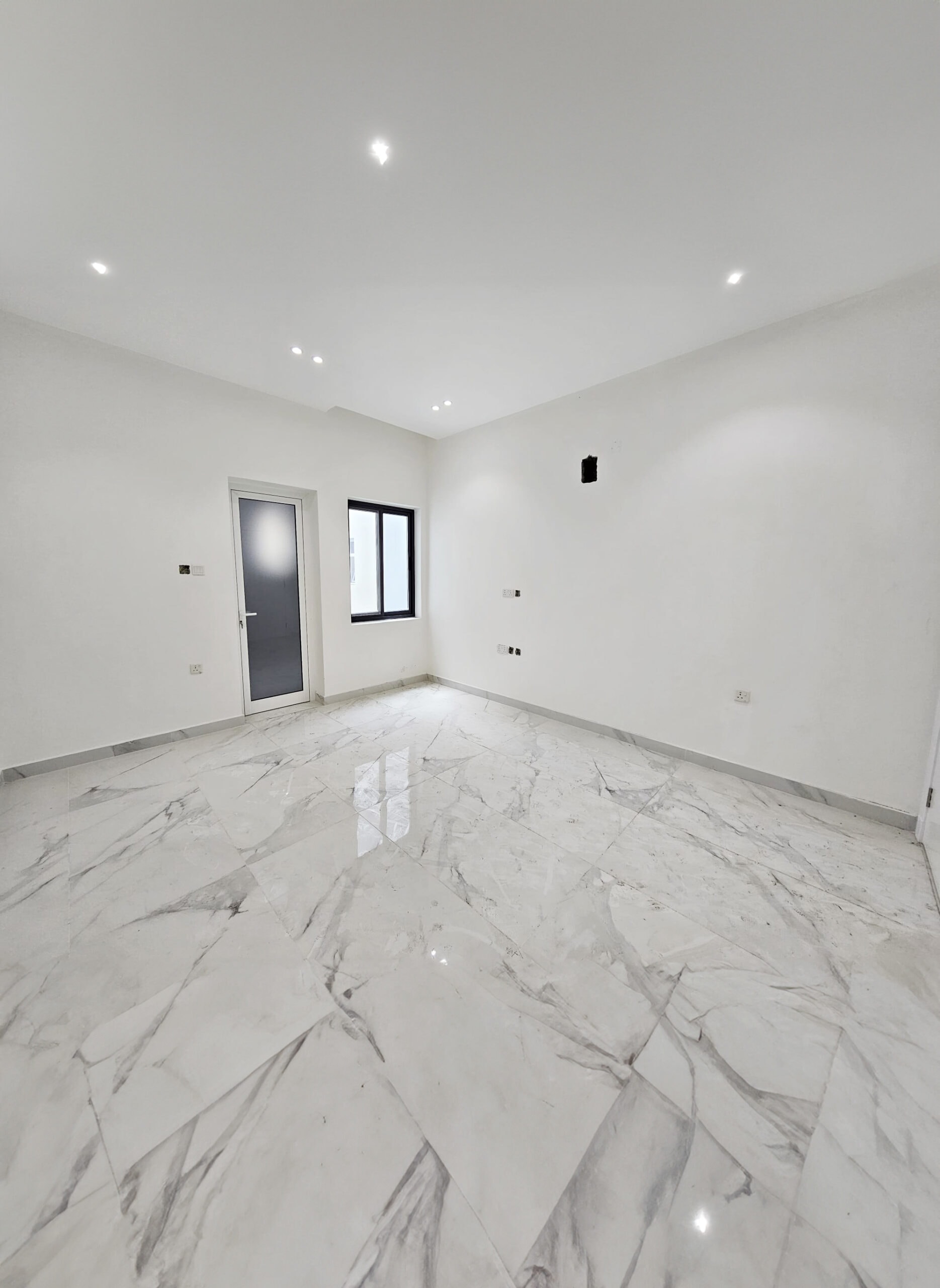 This spacious, empty room with white walls and a glossy marble floor is part of a luxurious villa. The room features a window, a door, and overhead recessed lighting, all designed to accentuate the elegance of this unique villa for sale.