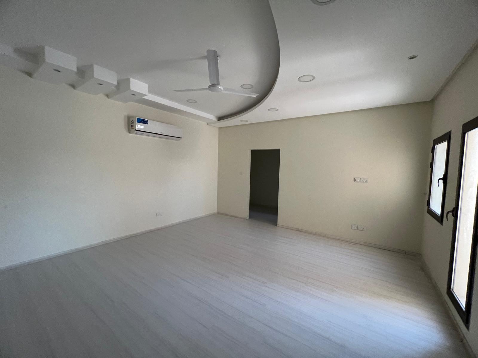 A beautiful villa for rent in Sanad features a vacant room with light-colored flooring, white walls, a ceiling fan, a wall-mounted air conditioner, and recessed lighting. Two windows on the right wall and a door leading to another room complete this serene space.