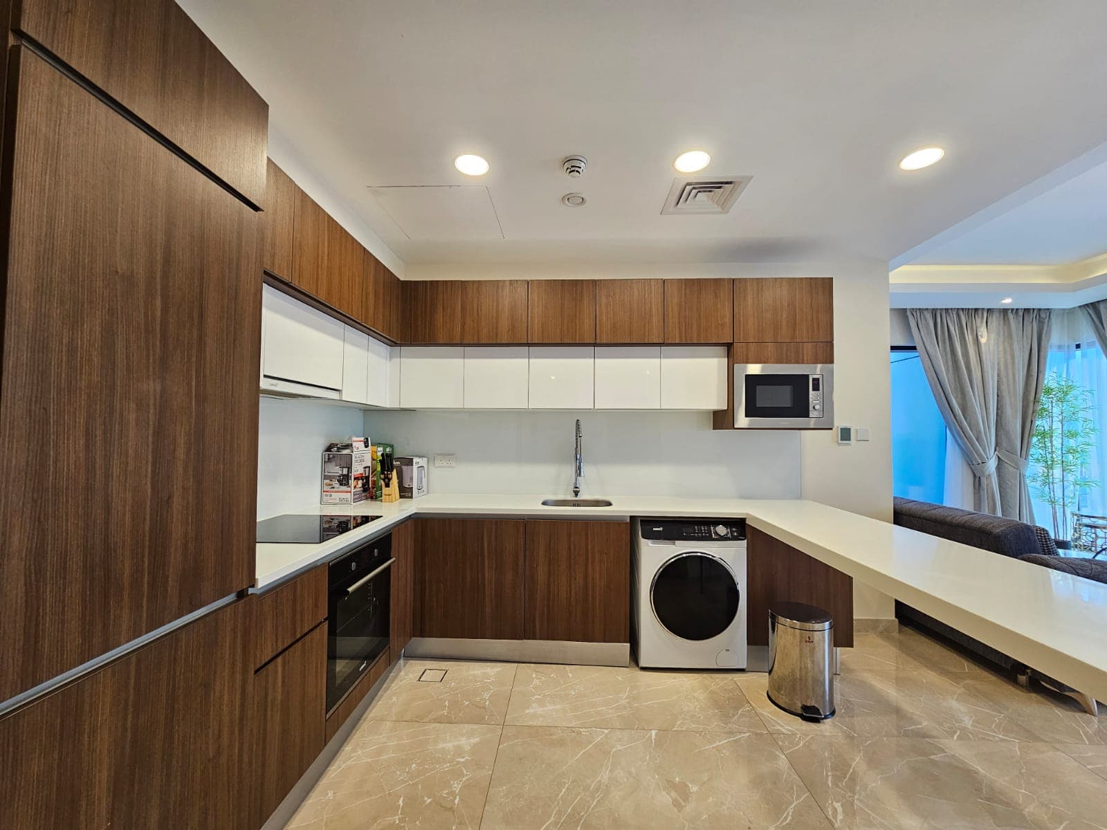 Modern kitchen with wooden cabinets, built-in appliances, and marble flooring, featuring a view of a curtain-covered window.