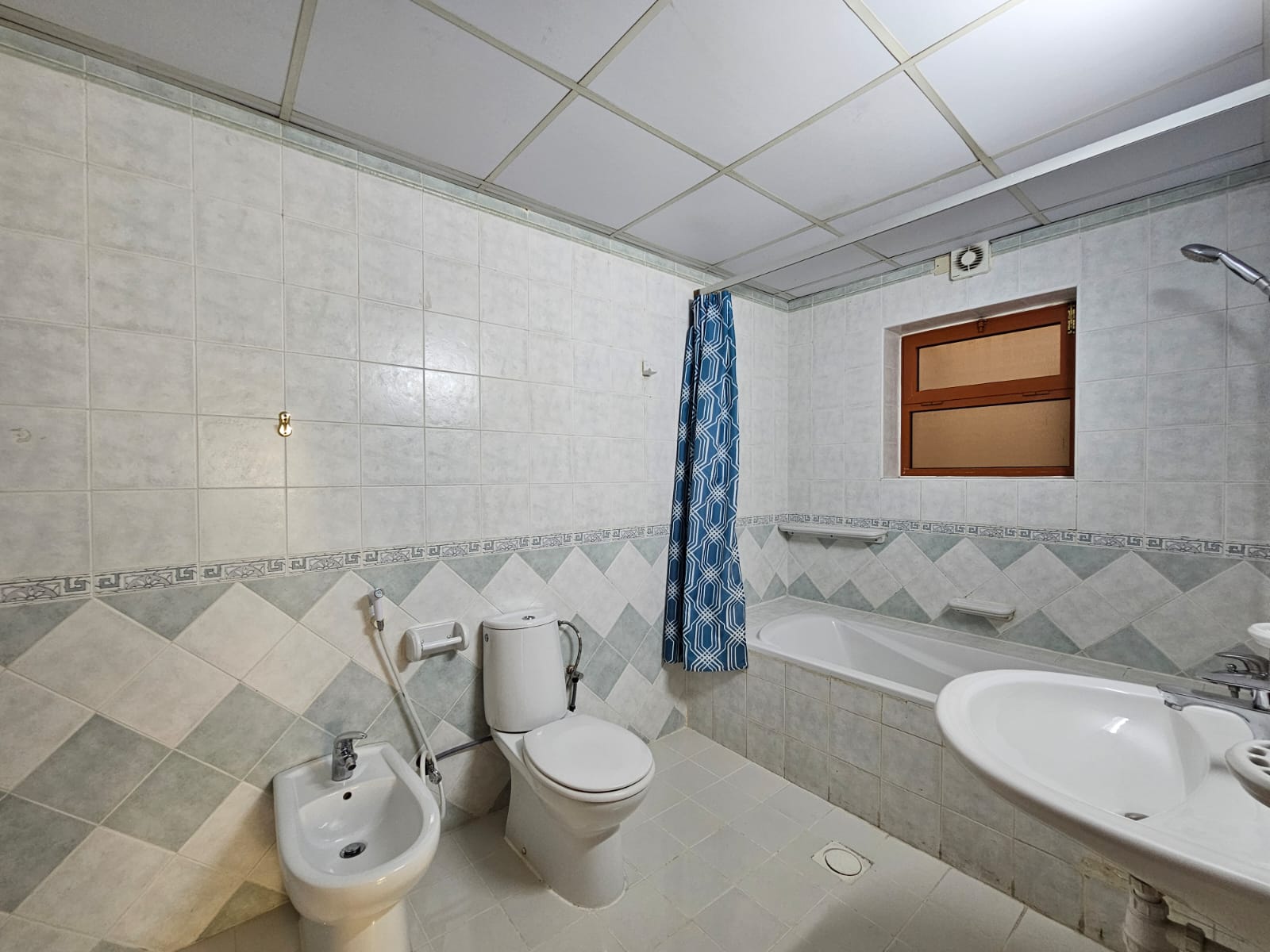 A small, tiled bathroom in a Juffair Area flat featuring a bathtub with a blue shower curtain, a toilet, bidet, sink, and a wooden cabinet above the sink.