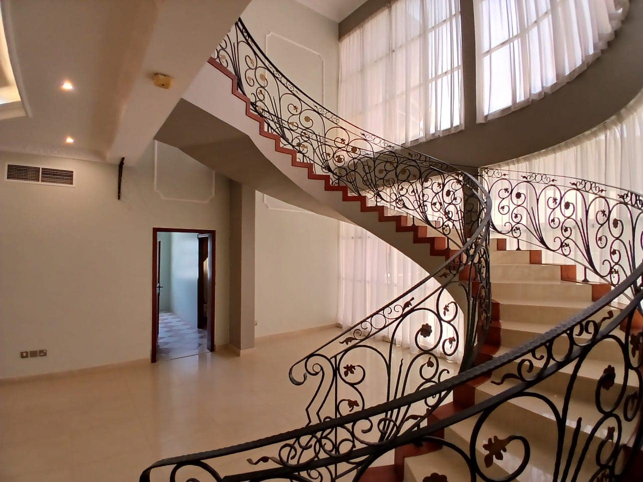 A spacious interior with a curved, ornate iron staircase. Large windows with sheer curtains allow ample natural light. An open doorway and tiled floor are visible on the left, perfect for a commercial villa available for rent.