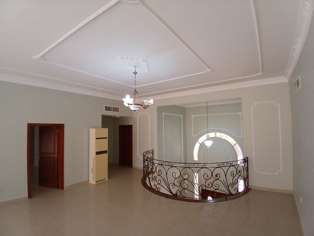 A spacious, unfurnished room in this commercial villa features a chandelier, ornate ceiling moldings, and a spiral staircase leading to a lower level. The room has beige walls and tile flooring, ideal for rent as an elegant office or boutique space.