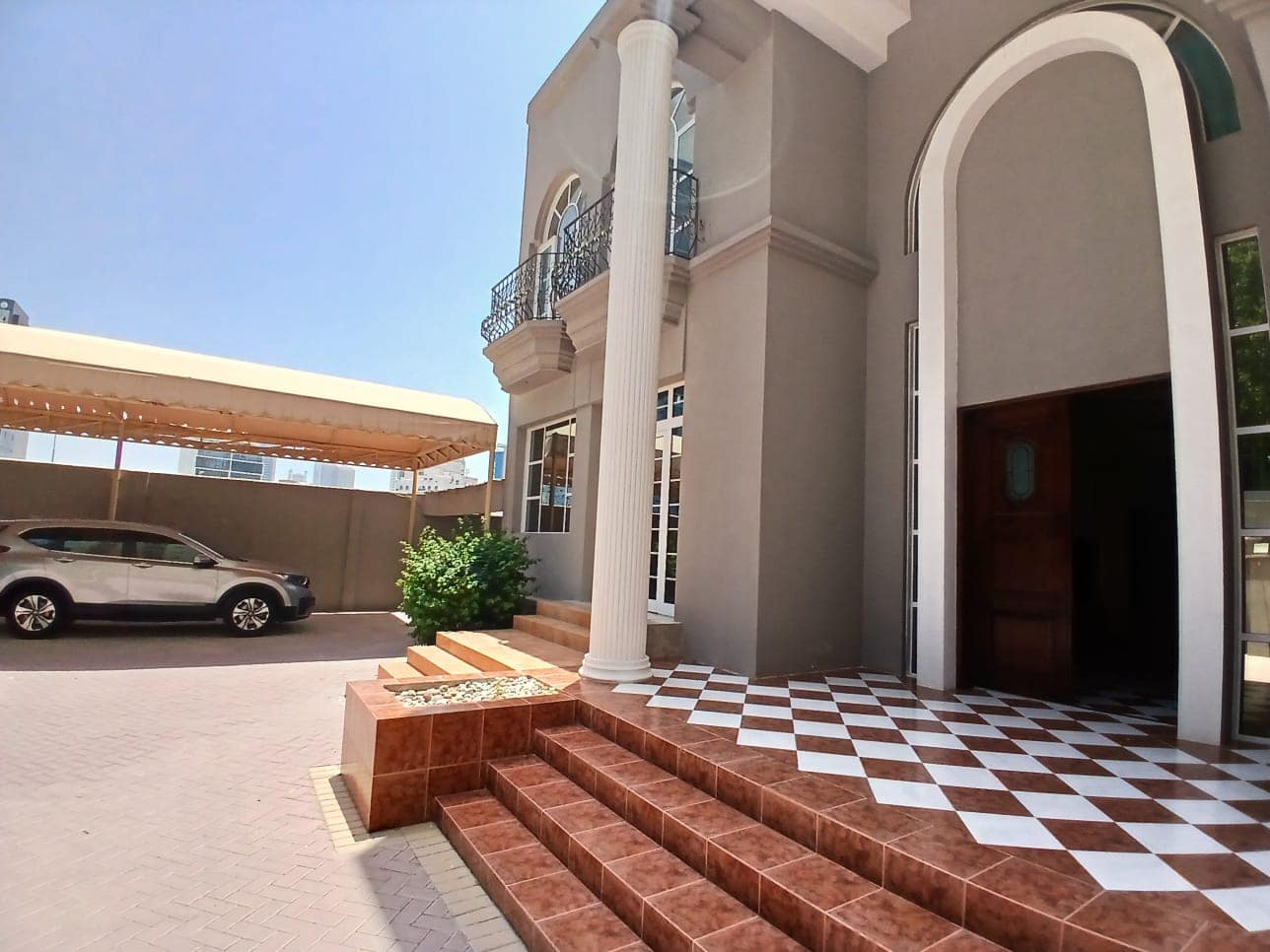 A modern commercial villa with gray walls and white columns. The entrance features a brown tiled staircase, ideal for professional settings. A car is parked under a beige canopy to the left, making it convenient for tenants and visitors alike. Perfect for rent!