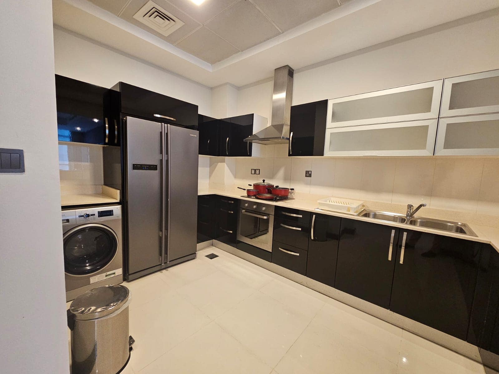 Modern kitchen with black cabinets, stainless steel fridge, oven, washing machine, and hood. Includes a double sink with a faucet and a dish rack. White tiled floors and walls with overhead lighting.