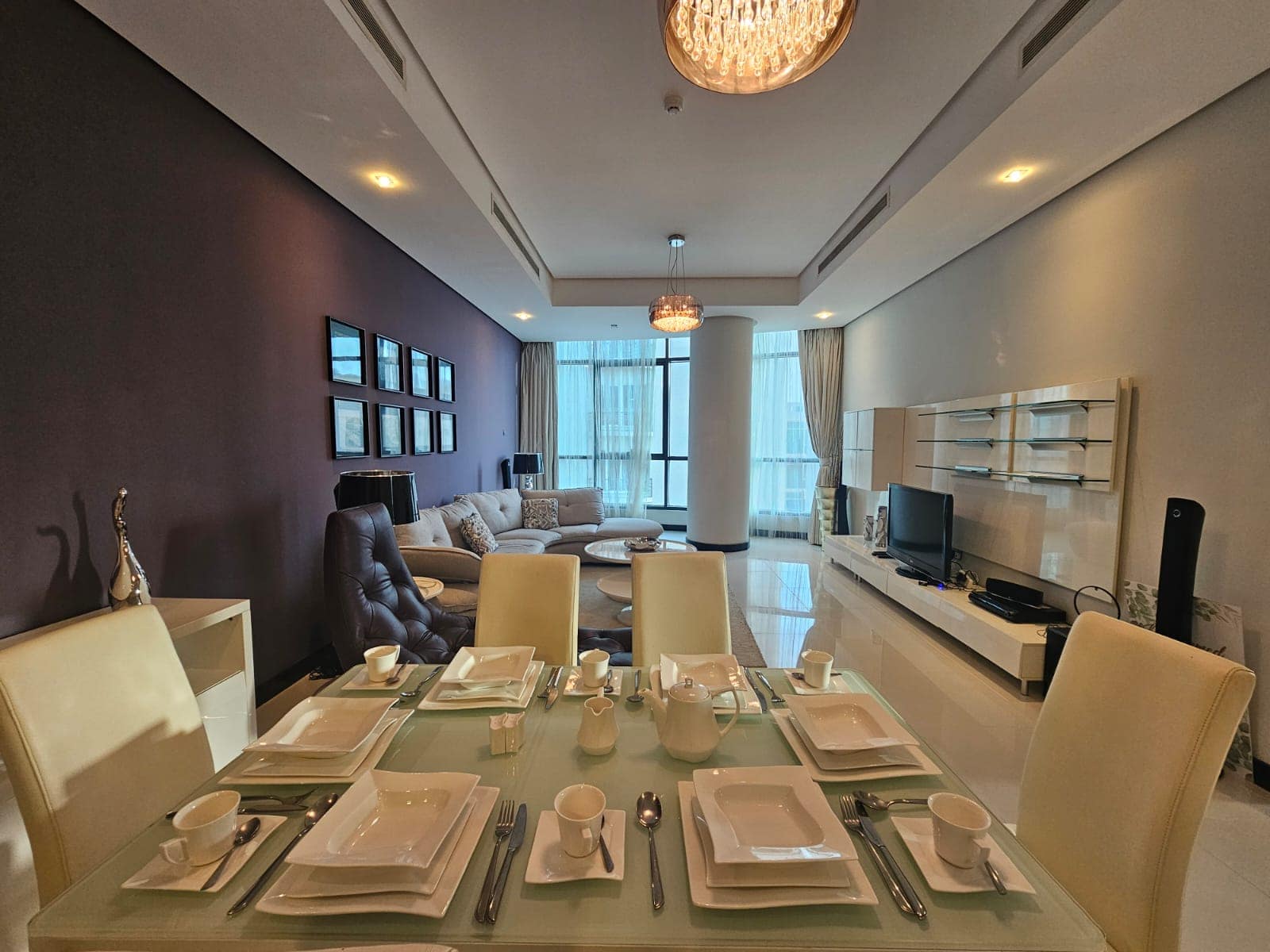 A modern, well-lit living and dining area with a glass dining table set for six, contemporary decor, a chandelier, a wall-mounted TV, and large windows covered with sheer curtains.