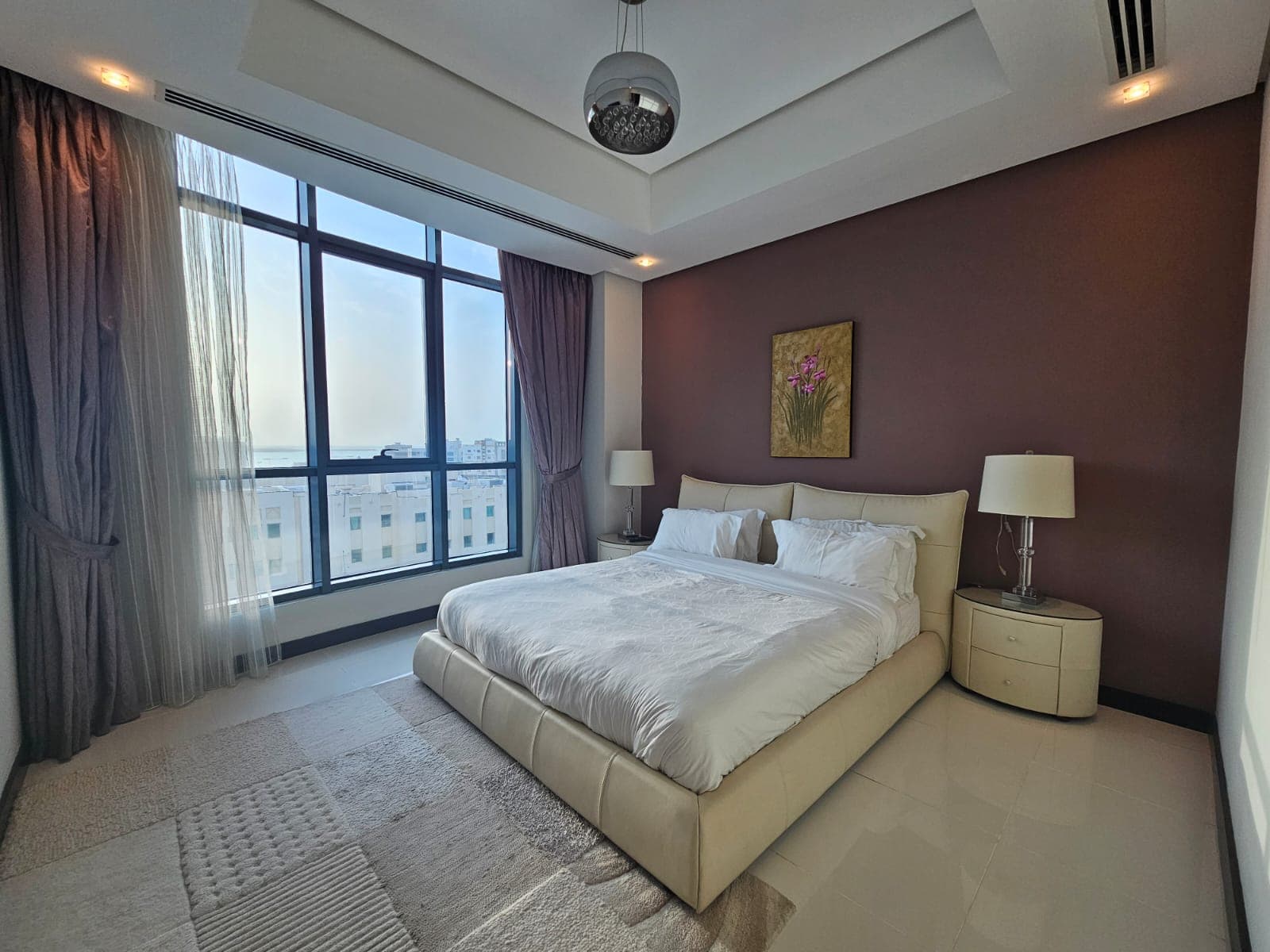 A modern bedroom with a large bed, white linens, two bedside tables with lamps, and a window with sheer curtains, showcasing a view of buildings outside. The walls are painted in neutral tones.